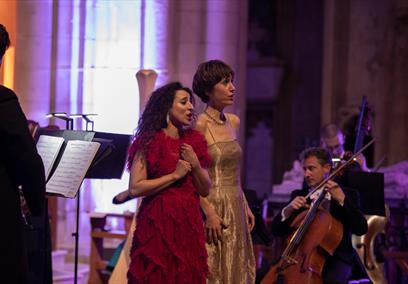 Two women singing together on stage with a chamber orchestra around them