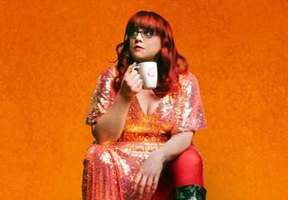 Woman with red hair wearing a glittery dress sat on an orange box against an orange backdrop holding a mug