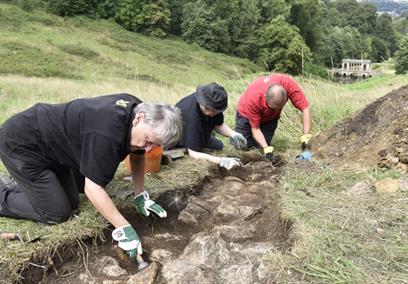 Volunteers digging in the Pasture uncovering stones from the Bason of Water with a view of Prior Park behind.