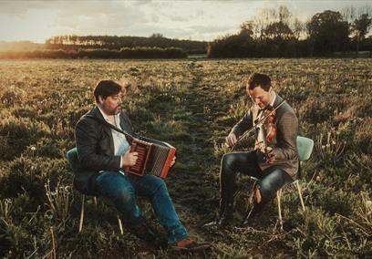 Spiers & Boden sitting in an empty field at sunset playing their instruments
