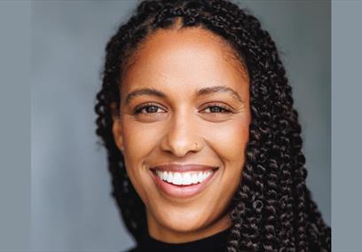 Tash Thomas smiles at the camera. She is a woman of colour with long braided hair.