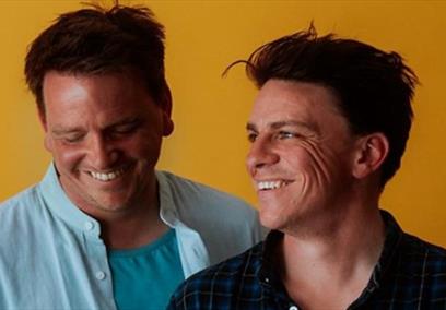 Two white men smiling and laughing whilst looking down, in front of a bright orange