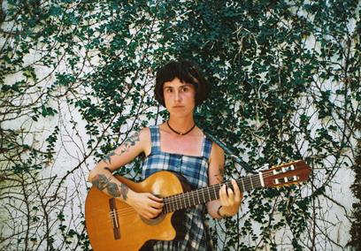 Maddi O'Brien playing a guitar in front of an ivy covered wall.