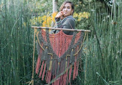A woman standing in tall grass holding a macrame tapestry