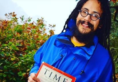 Rastafari historian Ras Benji stands in front of a hedge holding a copy of Time magazine. He has long dreadlocks and is smiling.