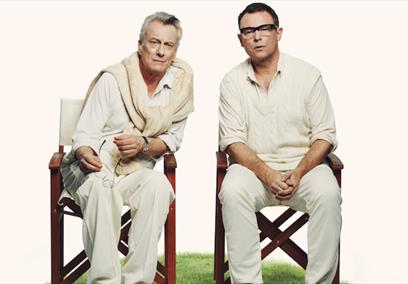 Two men sat on fold up chairs on a cricket pitch