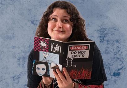 Tiff gazes whimsically at the camera with a diary covered in stickers in her hands. She wears a black t-shirt with long red and grey striped sleeves.