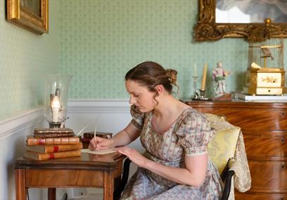 A woman in Regency clothing writing at a writing desk