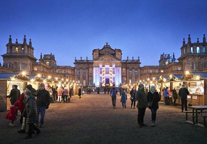 People walk amongst brightly lit chalets in front of Blenheim Palace
