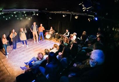 The cast of This Is Your Musical are performing on stage with two musicians and an audience is visible looking on at the performance.
