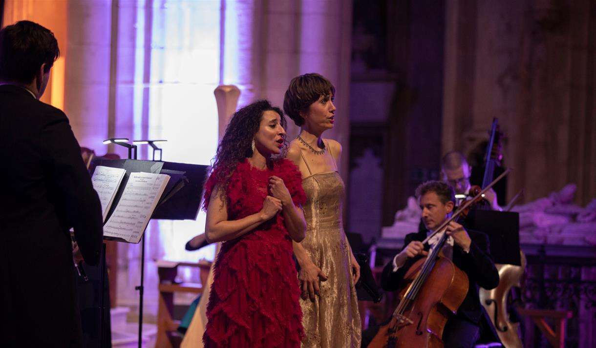 Two women singing together on stage with a chamber orchestra around them