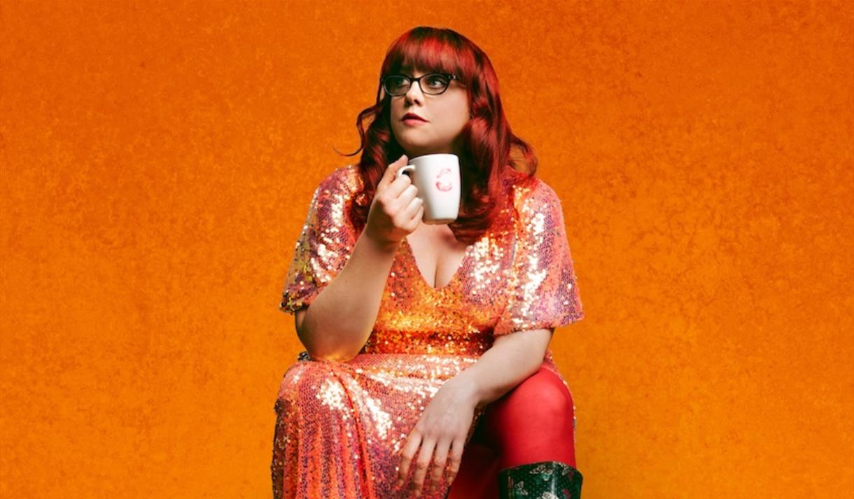 Woman with red hair wearing a glittery dress sat on an orange box against an orange backdrop holding a mug