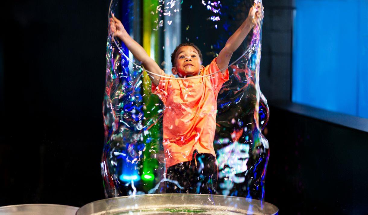 A small boy wearing an orange t shirt creating a large bubble
