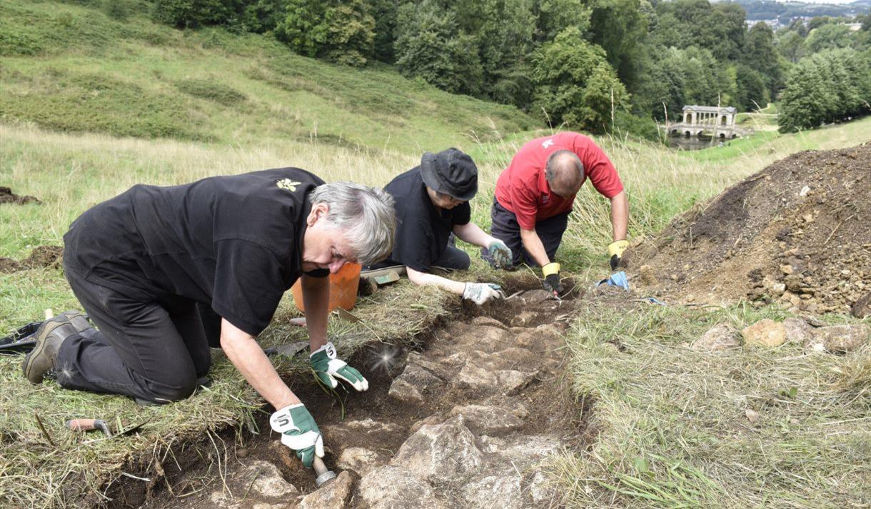 Volunteers digging in the Pasture uncovering stones from the Bason of Water with a view of Prior Park behind.