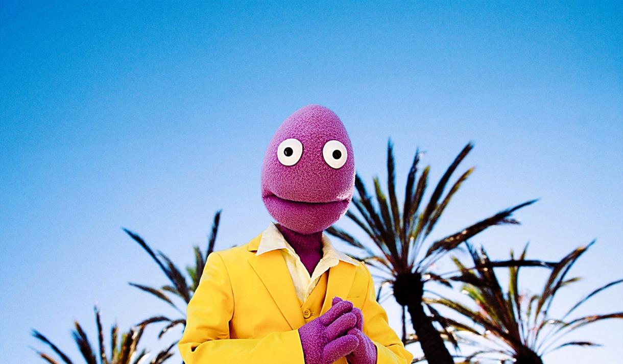 Randy Feltface in a yellow suit standing against a sunny background.