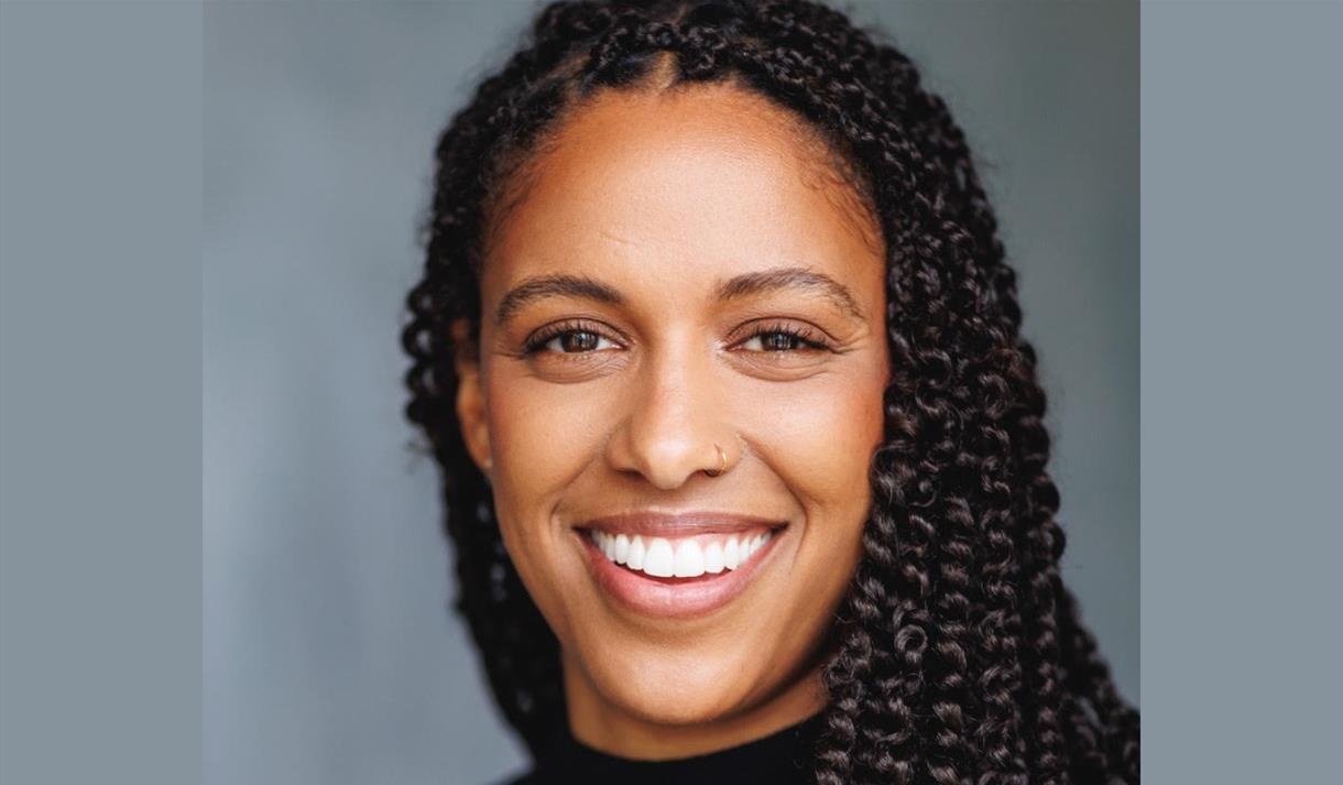 Tash Thomas smiles at the camera. She is a woman of colour with long braided hair.
