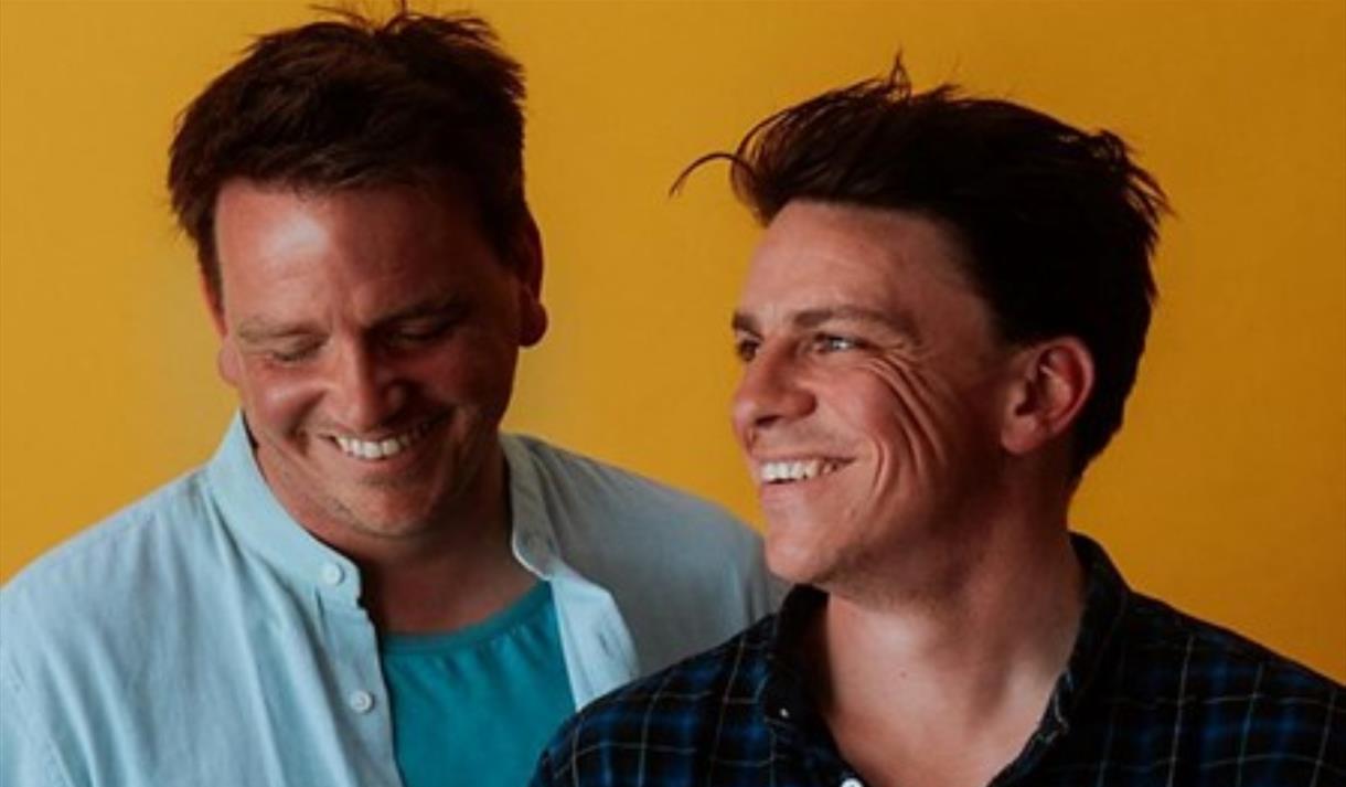 Two white men smiling and laughing whilst looking down, in front of a bright orange