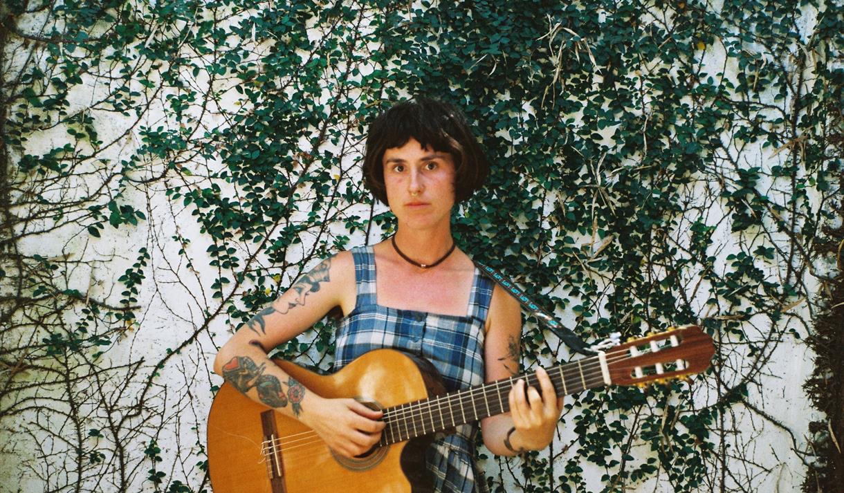 Maddi O'Brien playing a guitar in front of an ivy covered wall.