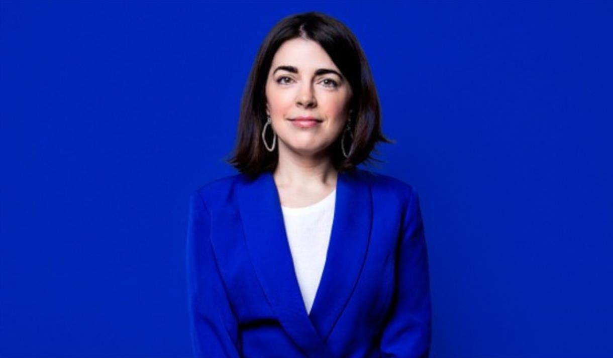 Rosie Holt in a blue suit sat infront of a blue background