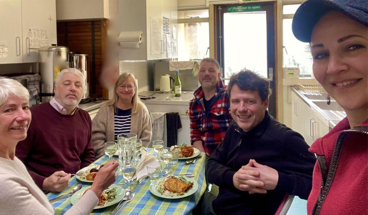 A group people sat at a table eating food with a women in a pink jacket taking their picture