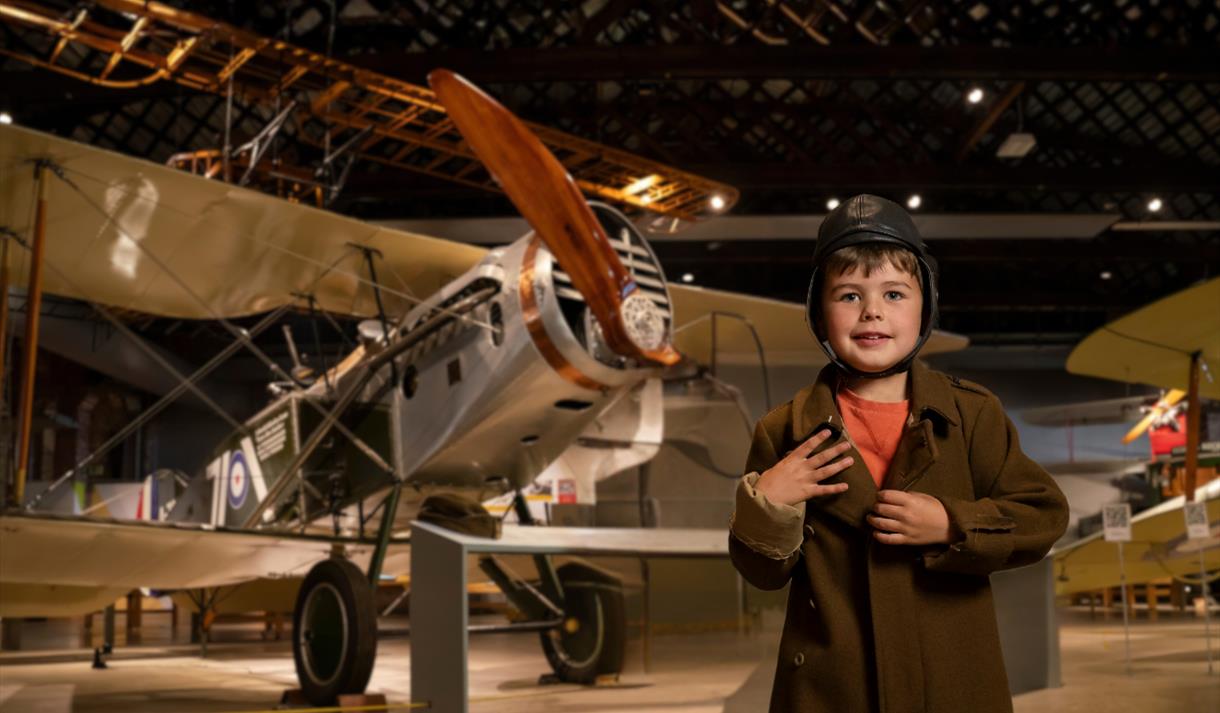A small boy wearing a brown vintage flying jacket and helmet, poses in front of a boxkite plane (a wooden plane) in a museum exhibit