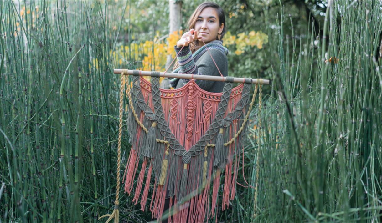 A woman standing in tall grass holding a macrame tapestry