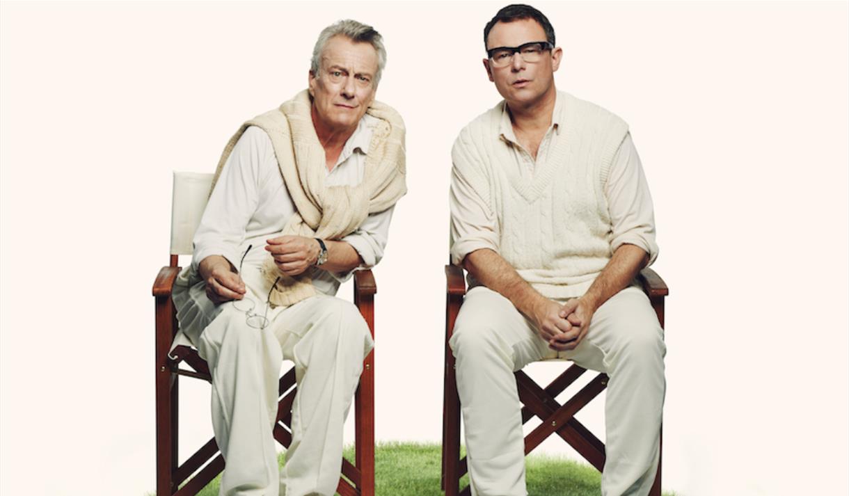 Two men sat on fold up chairs on a cricket pitch
