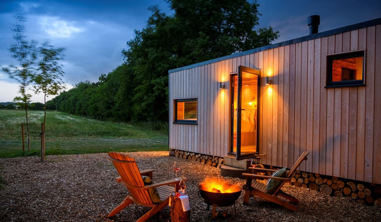 A photo of one of the cabins at dusk, showing the countryside view in the background, the glow of the firepit with 2 wooden armchairs around it and a