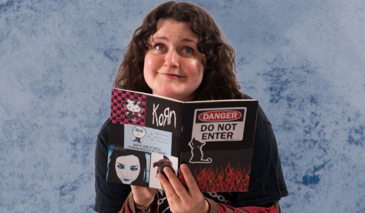 Tiff gazes whimsically at the camera with a diary covered in stickers in her hands. She wears a black t-shirt with long red and grey striped sleeves.