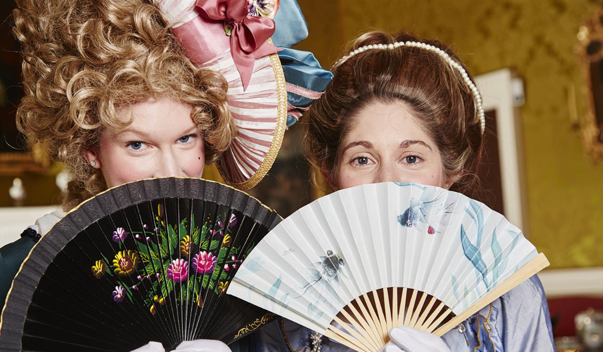 Two women dressed in Georgian outfits holding fans in front of their faces