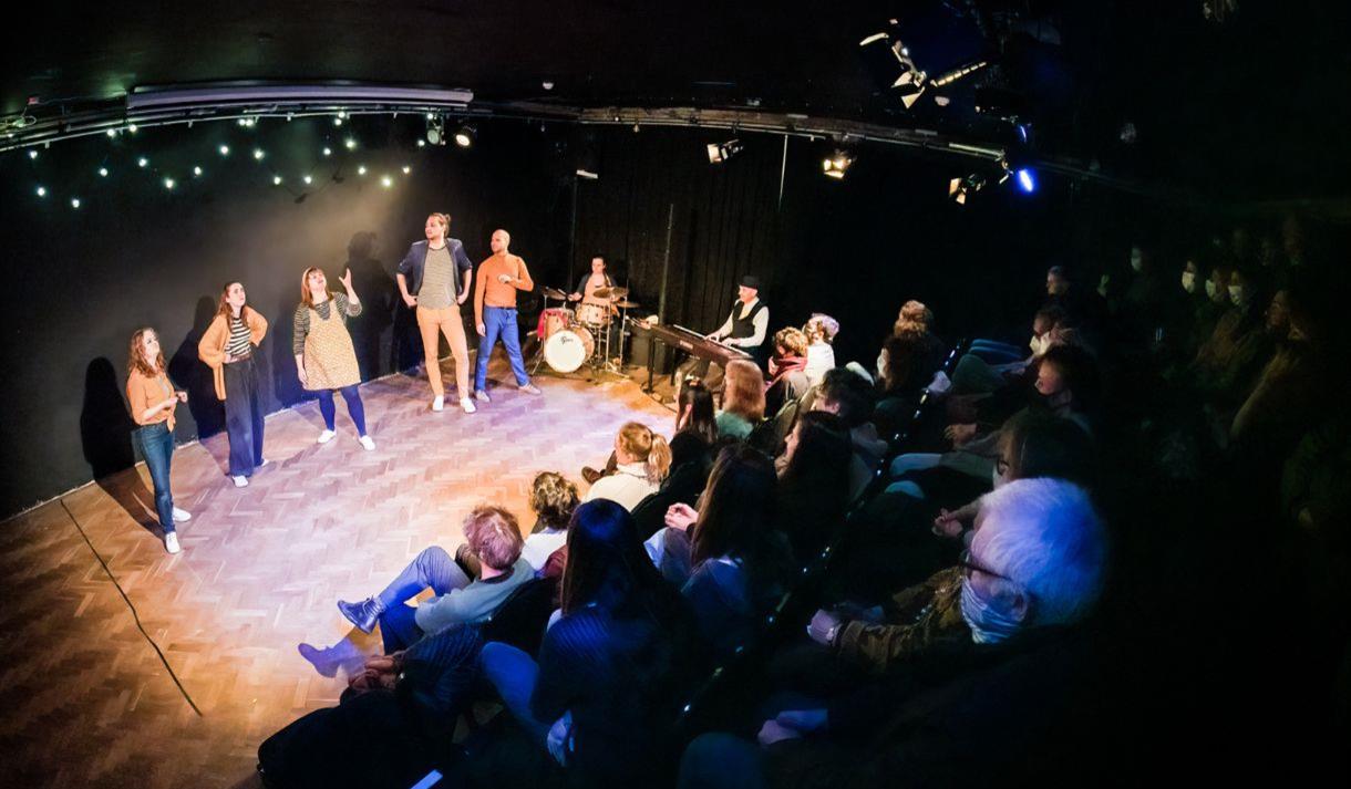 The cast of This Is Your Musical are performing on stage with two musicians and an audience is visible looking on at the performance.
