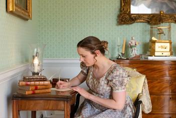 A woman in Regency clothing writing at a writing desk