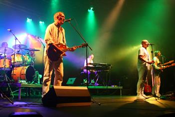 A photo of G2 Definitive Genesis performing on a stage. They are all wearing white clothing. Green, red, white and purple lights illuminate the stage.