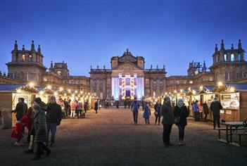 People walk amongst brightly lit chalets in front of Blenheim Palace