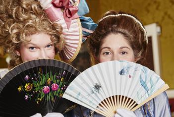 Two women dressed in Georgian outfits holding fans in front of their faces