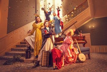 A group of women dressed in period costume. Three are at the bottom of a staircase grouped around a piano, while the other two are on the staircase, w