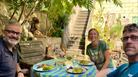 A group of three people sat around a sunny table in an attractive courtyard with white wine and plates of food.
