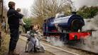 A family of four can be seen from behind waving at a blue steam train from the station platform.