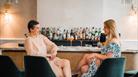 Two women sitting at a bar smiling at each other with cocktails behind them