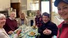 A group people sat at a table eating food with a women in a pink jacket taking their picture