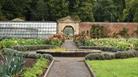 View across a garden with a path and flower bed in the middle with the path leading towards an arch attached to a greenhouse