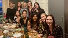 A group of ten girls smiling and toasting along a long wooden table with empty bowls and wine glasses on the table.