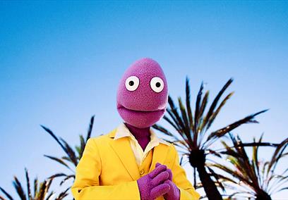 Randy Feltface in a yellow suit standing against a sunny background.
