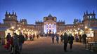 People walk amongst brightly lit chalets in front of Blenheim Palace