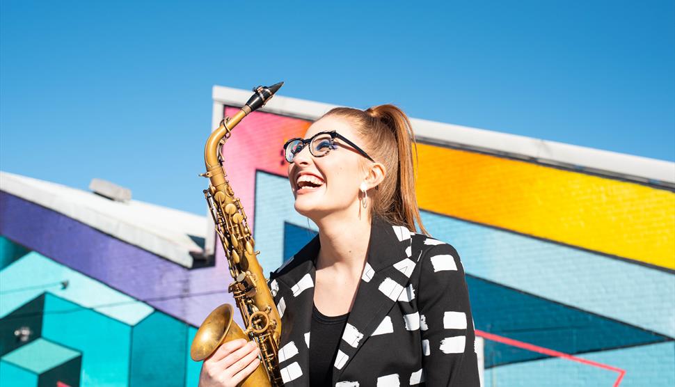 A ginger haired woman wearing glasses smiles while holding a saxophone. The wall behind her is blue and colourful.
