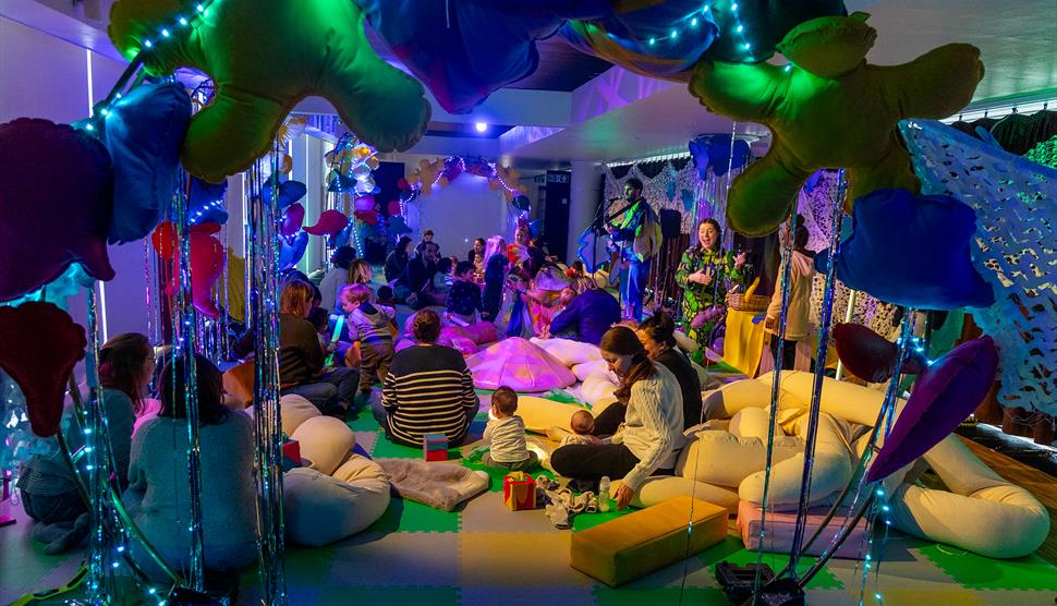 A group of children and parents sit in a colourful room with cushions and balloons. On one side, a band plays music to the parents and children.