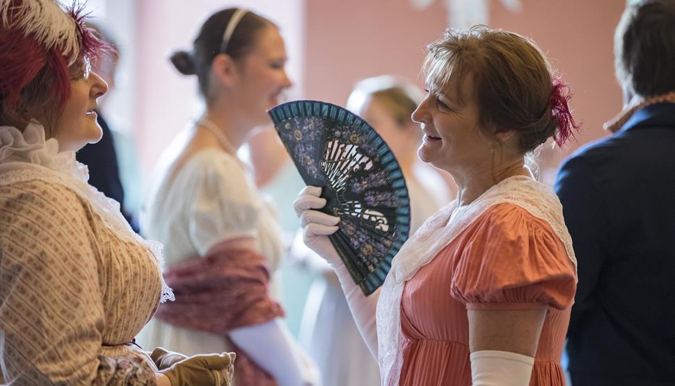 Ladies in Regency costume chat at an event. A lady on the right uses a black fan.