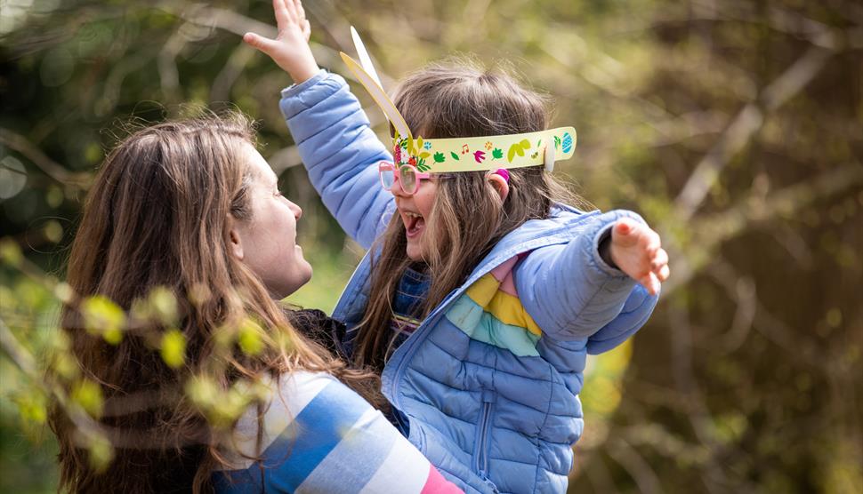 A parent lifts their child up. The child wears bunny ears and a lilac coat and has their arms up in the air.