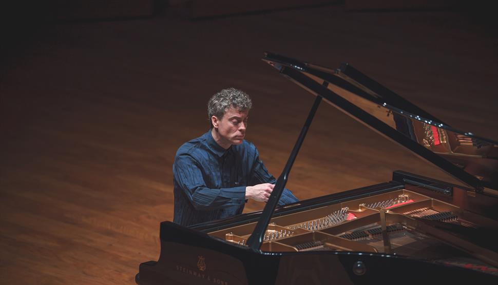 Paul Lewis is playing the piano in a dimly lit concert hall.