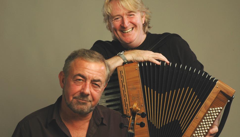Phil Cunningham is standing, holding his accordion and smiling, with Aly Bain sitting in front of him holding his fiddle.